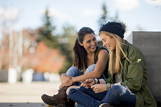 Two college age girl friends are sitting outside on a beautiful fall day. They are talking together before class.